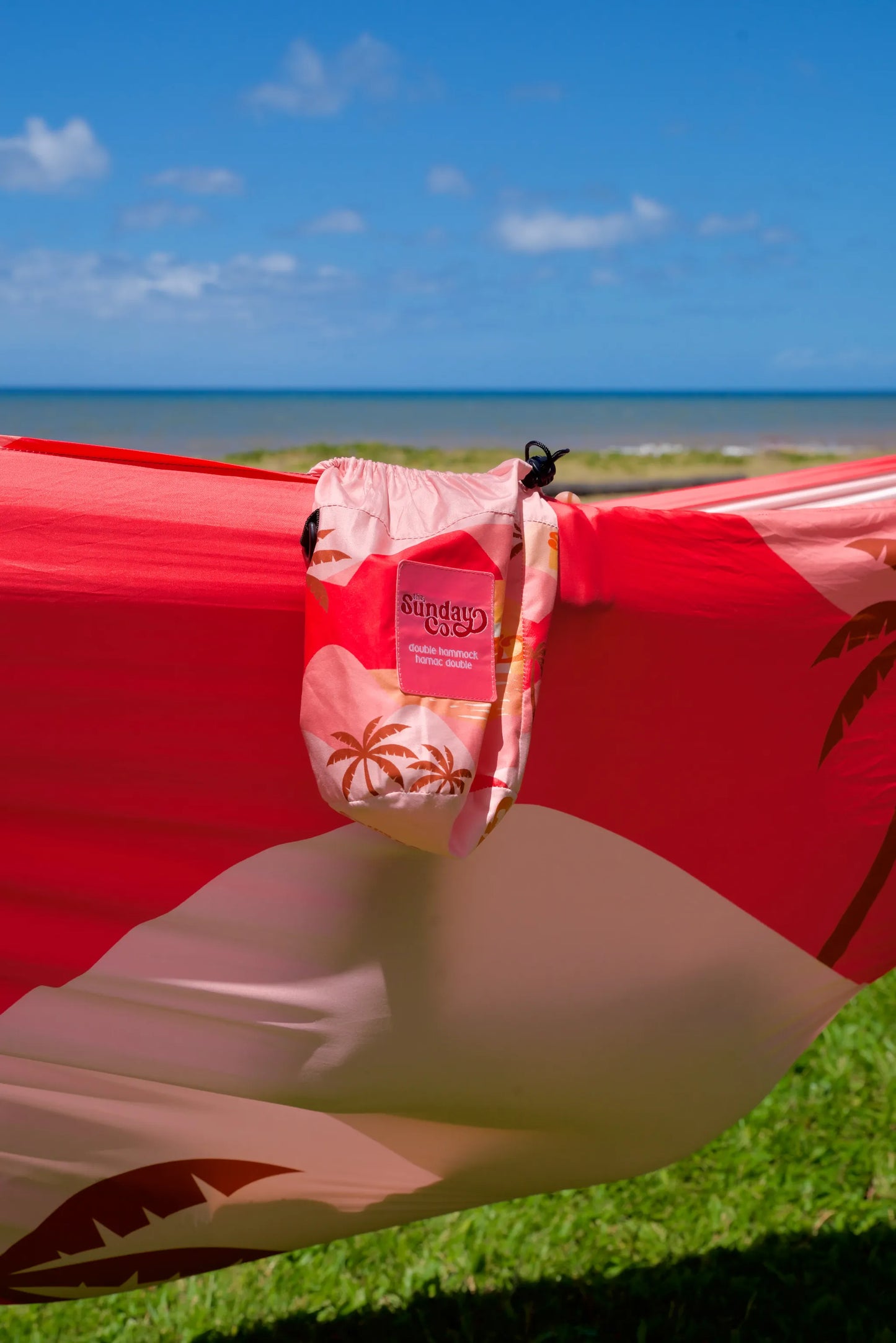 The Sunday Company Pink Sunets Hammock - Several shades of pink with dark rosy brown palm pattern throughout, cute matching carry bag attached. Hammock is strung up on a grassy knoll near the ocean.