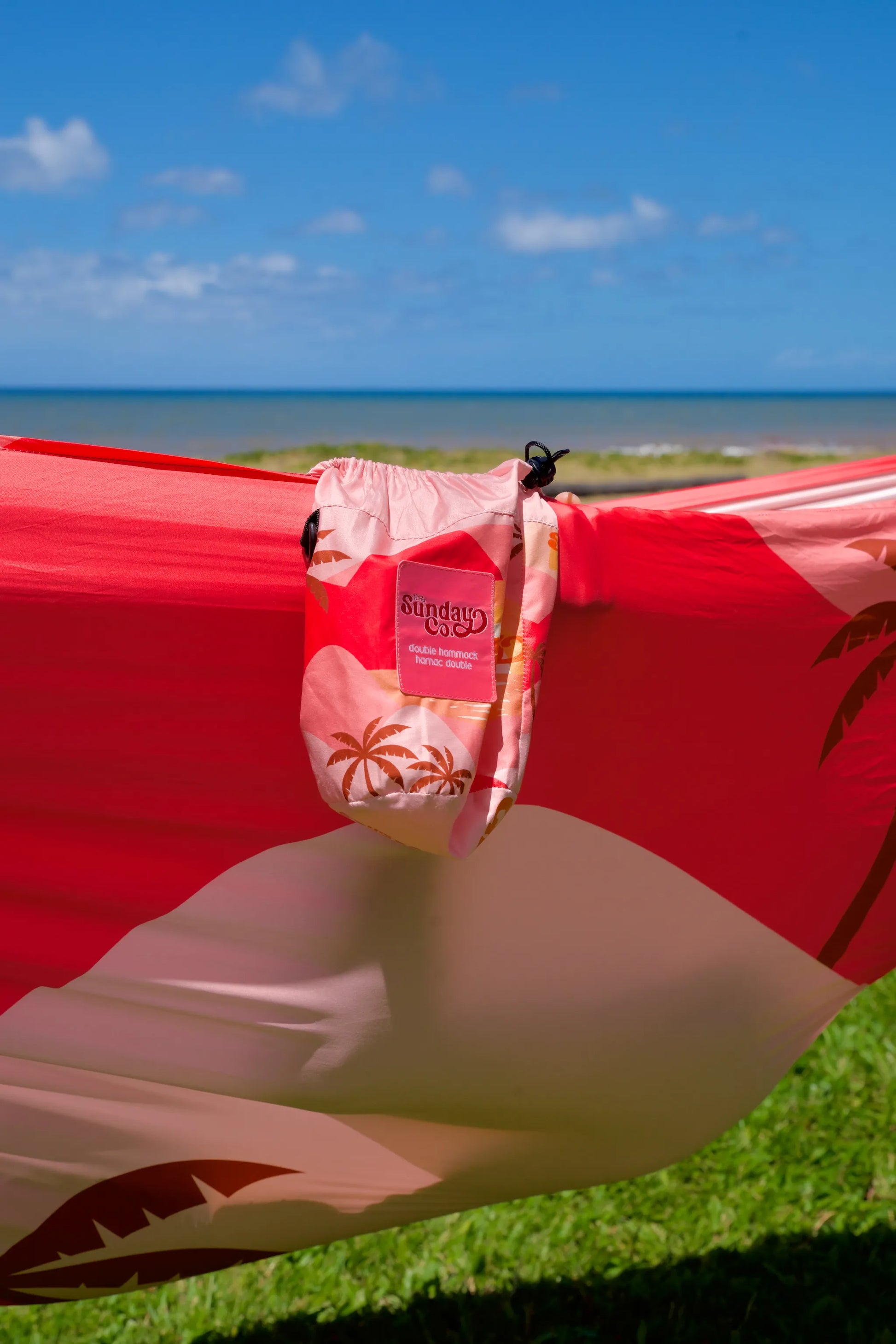The Sunday Company Pink Sunets Hammock - Several shades of pink with dark rosy brown palm pattern throughout, cute matching carry bag attached. Hammock is strung up on a grassy knoll near the ocean.