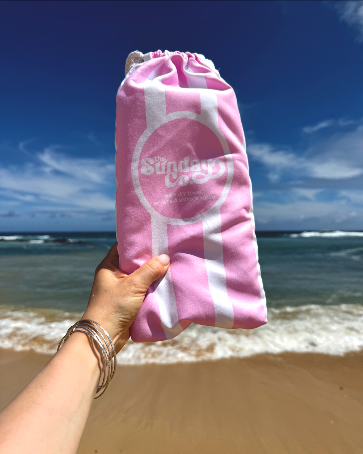 Pink drawstring bag with 'The Sunday Club' logo held by a hand on a beach.