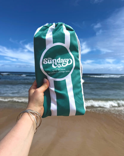 Green and white drawstring bag with 'The Sunday Co.' logo held by a hand on a beach.
