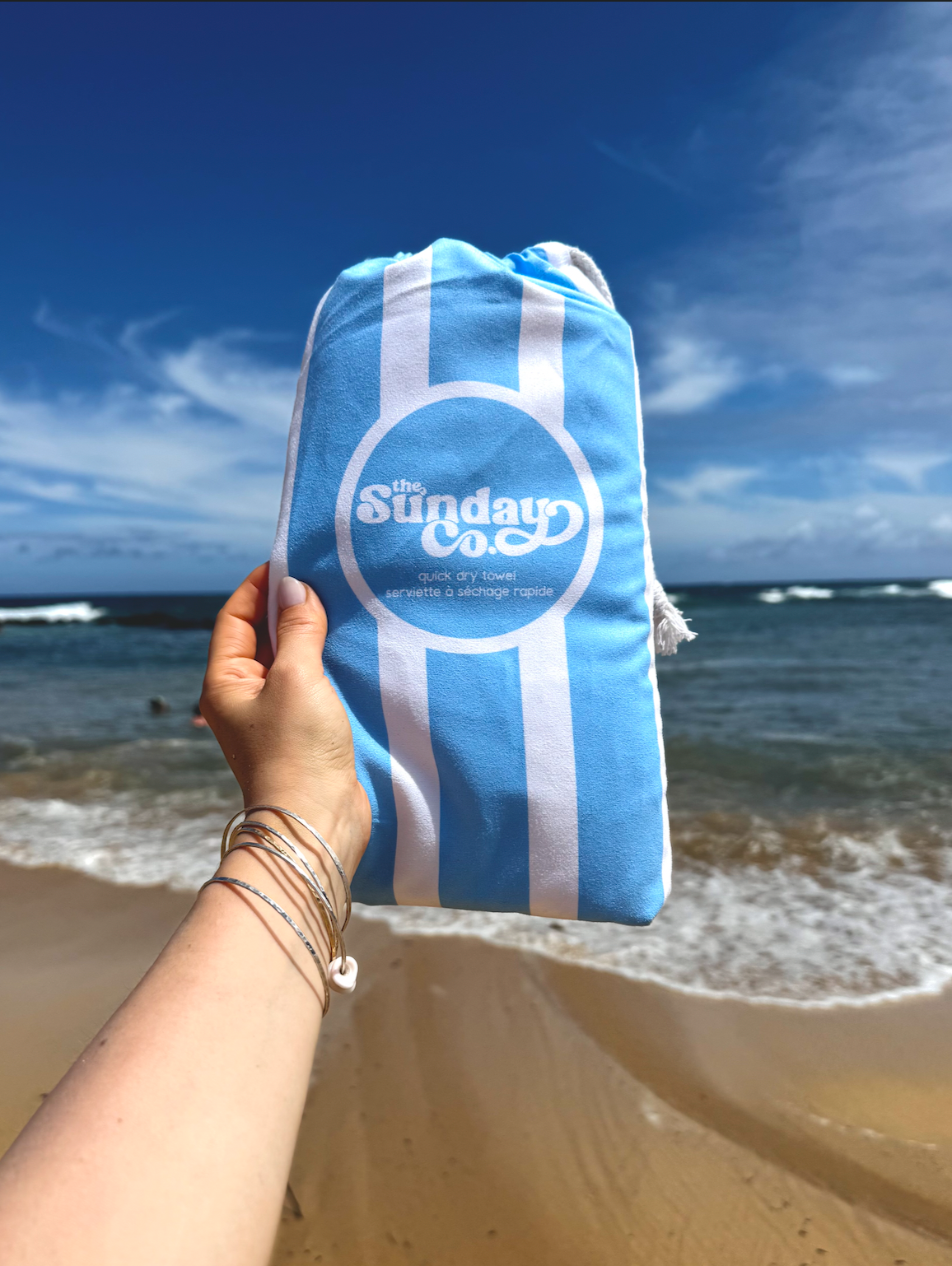 Hand holding a blue and white striped bag with 'The Sunday Co.' logo on a beach.