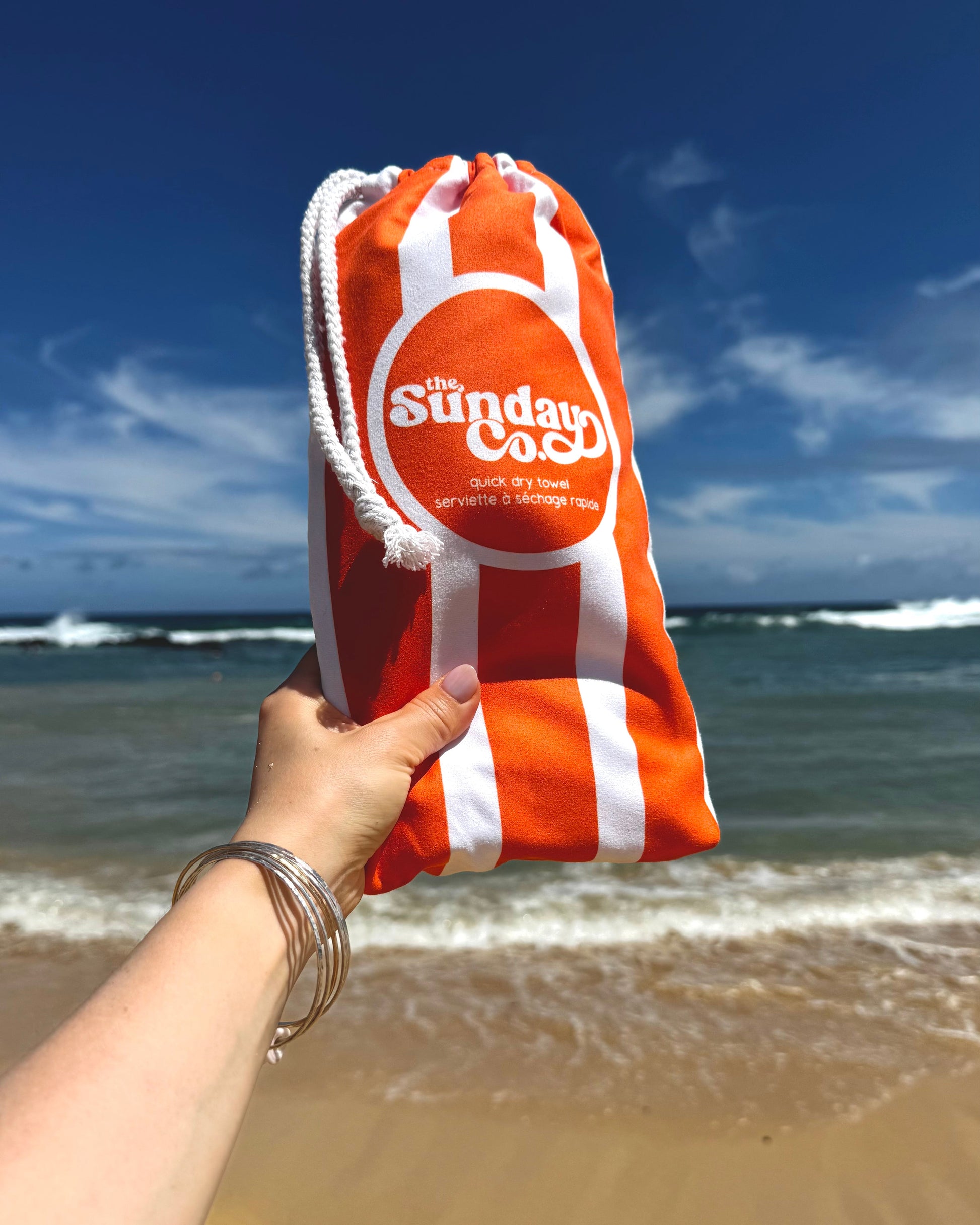 Orange and white bag with 'The Sunday Cook' logo held by a hand on a beach.